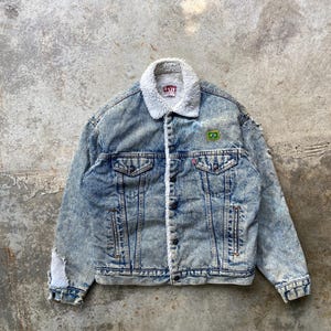 Könnte beinhalten: Hellblaue Jeansjacke mit weißem Kunstfellkragen und -futter. Die Jacke hat eine Knopfleiste, zwei Brusttaschen und einen kleinen brasilianischen Flaggenaufnäher. Das Levi's-Logo ist sichtbar.