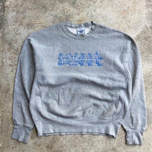 Op de afbeelding: Een grijze sweatshirt met het woord "BUMBLE" in blauwe blokletters op de borst. De sweatshirt heeft een ronde hals en geribde manchetten en zoom. Het Lee-logo is zichtbaar op de kraag.
