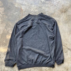 May include: A dark gray sweatshirt with long sleeves and a crew neck. The back of the sweatshirt is visible, showing the texture of the fabric. The cuffs and hem are a darker shade of gray.