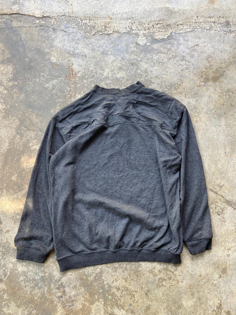 May include: A dark gray sweatshirt with long sleeves and a crew neck. The back of the sweatshirt is visible, showing the texture of the fabric. The cuffs and hem are a darker shade of gray.