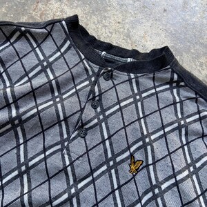 May include: A gray and white plaid Henley shirt with a black collar and button placket. The shirt features a small gold emblem on the chest. The pattern is a diamond grid design.