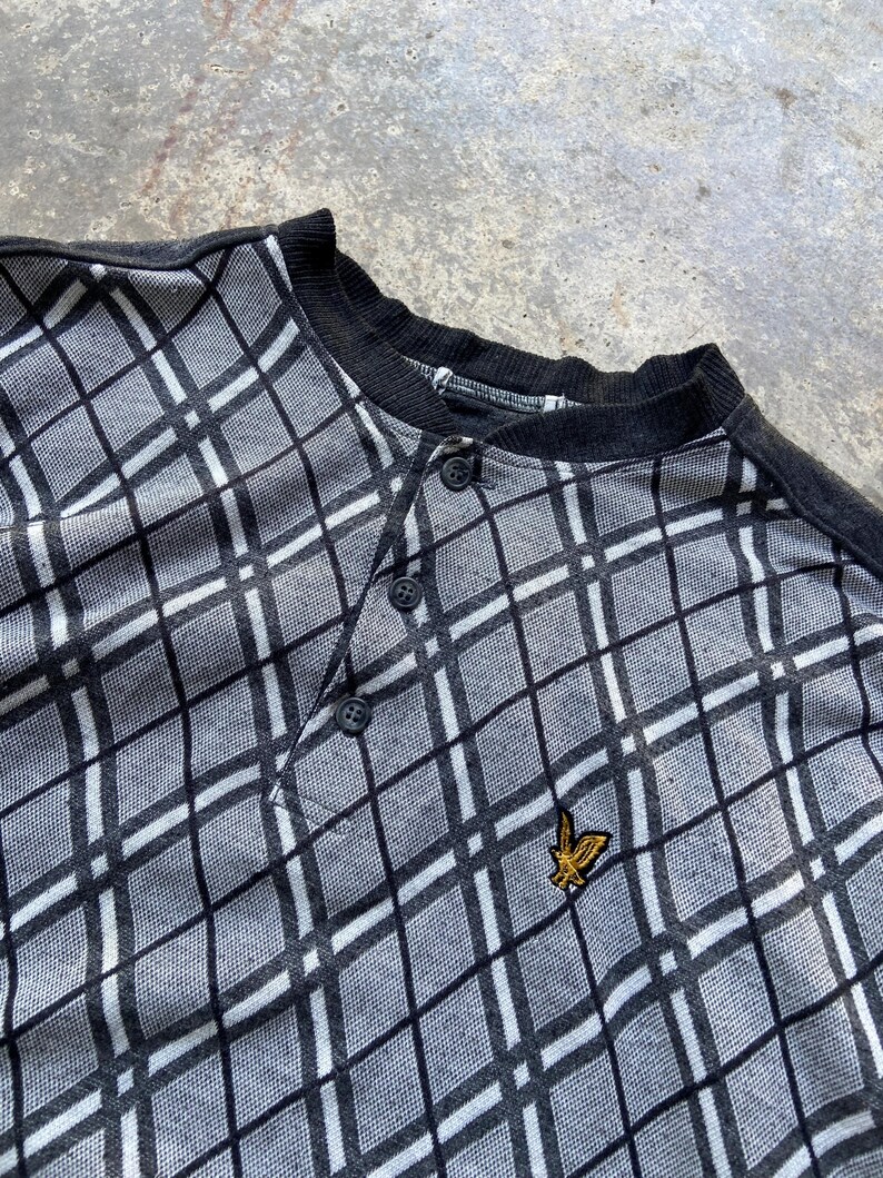 May include: A gray and white plaid Henley shirt with a black collar and button placket. The shirt features a small gold emblem on the chest. The pattern is a diamond grid design.