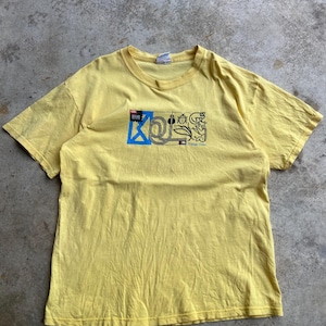 May include: A short-sleeved yellow t-shirt featuring a graphic print on the chest. The design includes a blue square, black line art of a beetle, a monkey, and a bird, along with the text "KOLS" and "Pura Vida" in a stylized black font.