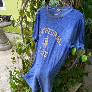 Puede incluir: Una camiseta azul descolorida con las palabras "CATHEDRAL SAINTS" en letras doradas. La camiseta está colgada de una percha de metal en medio de follaje verde. La camiseta parece desgastada, con un aspecto envejecido.