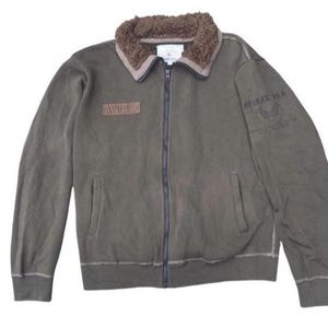 Könnte beinhalten: Olivgrüne Reißverschlussjacke mit braunem Kunstfellkragen. Die Jacke hat den Markennamen "AVIREX" auf einem Aufnäher und Text auf dem Ärmel. Sie hat zwei Vordertaschen und gerippte Bündchen und Saum.