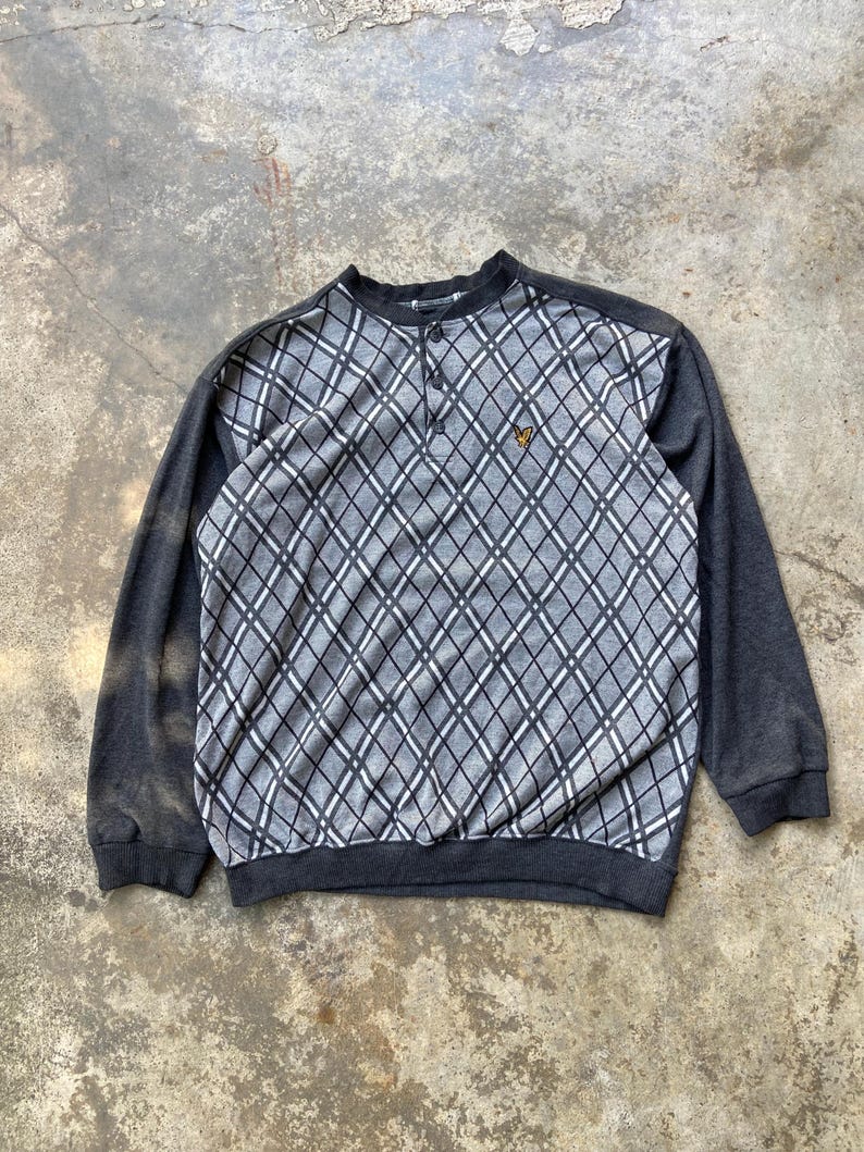 May include: A gray and black argyle patterned sweatshirt with long sleeves. The top portion features a diamond pattern with a button-up neckline and a small yellow logo. The sleeves and bottom hem are solid gray.
