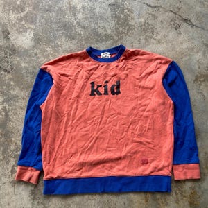 K&ouml;nnte beinhalten: Ein lang&auml;rmeliges Sweatshirt mit Rundhalsausschnitt, korallenfarbenem Rumpf sowie leuchtend blauen &Auml;rmeln, Kragen und Saum. Auf der Brust befindet sich der in schwarzen Buchstaben gedruckte Schriftzug "kid" und unten ein kleines Markenetikett.