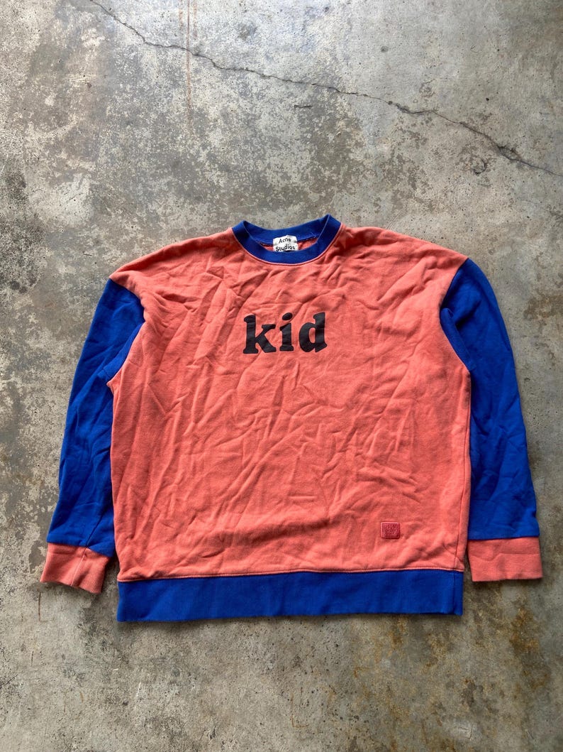 K&ouml;nnte beinhalten: Ein lang&auml;rmeliges Sweatshirt mit Rundhalsausschnitt, korallenfarbenem Rumpf sowie leuchtend blauen &Auml;rmeln, Kragen und Saum. Auf der Brust befindet sich der in schwarzen Buchstaben gedruckte Schriftzug "kid" und unten ein kleines Markenetikett.