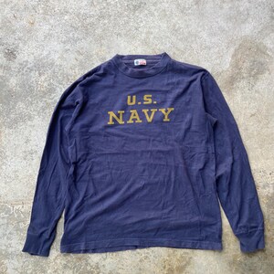 May include: Navy blue long-sleeve shirt with the text "U.S. NAVY" in gold lettering. The shirt has a crew neck and a vintage look. The fabric appears to be a soft, worn cotton.