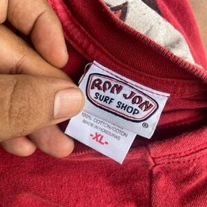 Puede incluir: Una camiseta roja con una etiqueta interior blanca en el cuello con el texto RON JON SURF SHOP en letras rojas y negras. La etiqueta tambi&eacute;n muestra 100% COTTON/COTON, MADE IN HONDURAS y la talla XL en texto rojo sobre fondo blanco.