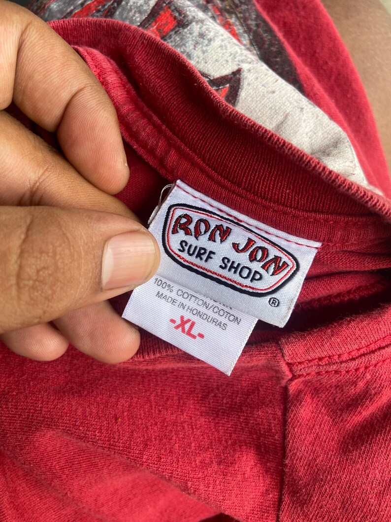 Puede incluir: Una camiseta roja con una etiqueta interior blanca en el cuello con el texto RON JON SURF SHOP en letras rojas y negras. La etiqueta tambi&eacute;n muestra 100% COTTON/COTON, MADE IN HONDURAS y la talla XL en texto rojo sobre fondo blanco.