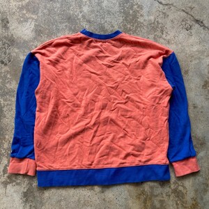 K&ouml;nnte beinhalten: Ein lang&auml;rmeliges Sweatshirt mit einem korallenfarbenen Hauptteil und einem Rundhalsausschnitt. Die &Auml;rmel sind einfarbig blau mit korallenfarbenen B&uuml;ndchen, und der untere Saum ist mit einem breiten blauen Band versehen, was ein Color-Block-Design ergibt.