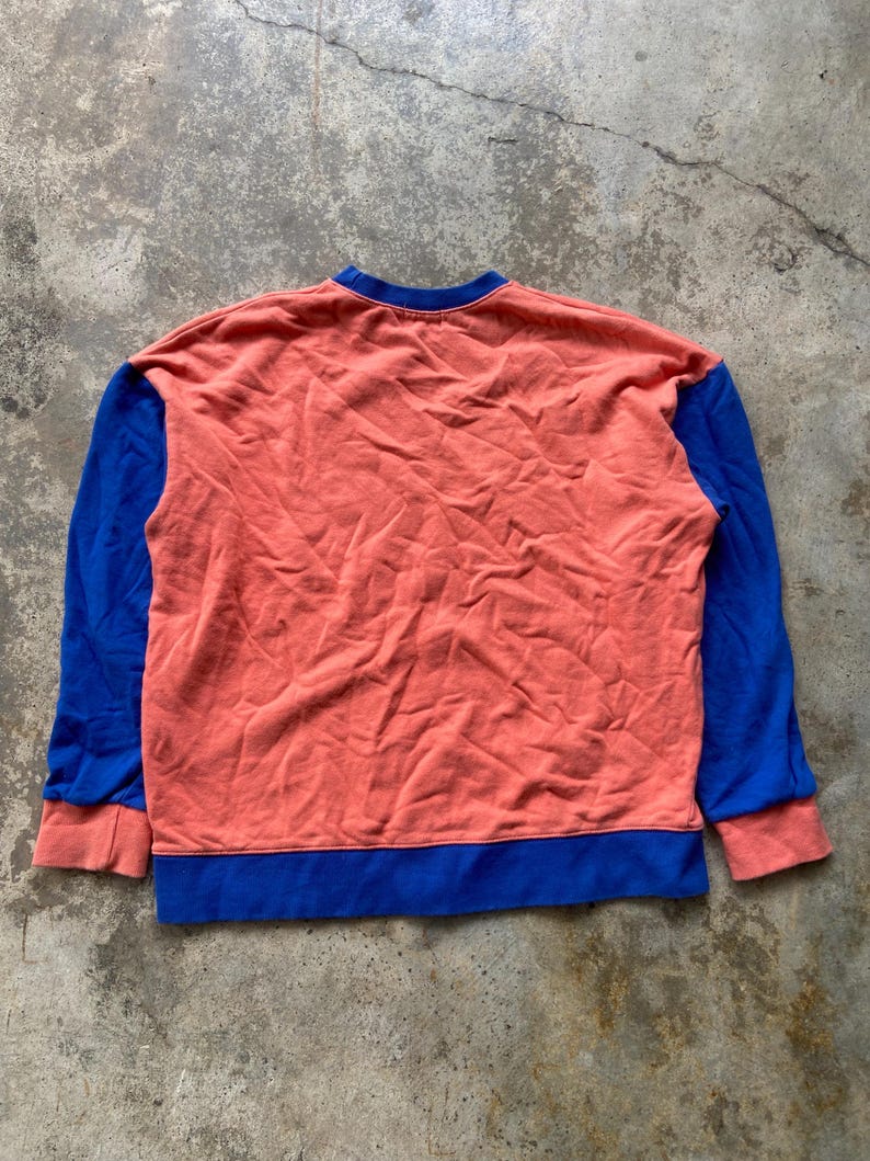 K&ouml;nnte beinhalten: Ein lang&auml;rmeliges Sweatshirt mit einem korallenfarbenen Hauptteil und einem Rundhalsausschnitt. Die &Auml;rmel sind einfarbig blau mit korallenfarbenen B&uuml;ndchen, und der untere Saum ist mit einem breiten blauen Band versehen, was ein Color-Block-Design ergibt.