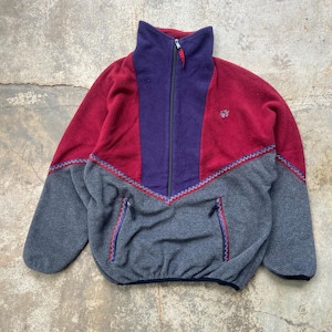 May include: A color-blocked fleece jacket in shades of red, purple, and gray. The jacket features a high collar, a full-length zipper, and two front pockets. The design includes a decorative zigzag pattern and a small logo on the chest.