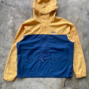 Puede incluir: Una chaqueta tipo pullover amarilla y azul con capucha, mangas largas y una solapa frontal que presenta un pequeño logotipo de la marca Patagonia en el pecho. La prenda incluye un cordón en la cintura y cierres de botones a presión al frente.