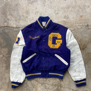 Könnte beinhalten: Eine blaue und weiße Vintage-Collegejacke. Auf der Vorderseite der Jacke ist der Name "Michael" aufgestickt. Sie hat einen großen gelben "G"-Aufnäher mit Flügeln und Sternen und die Nummer "2" auf dem linken Ärmel. Die Ärmel sind weiß.
