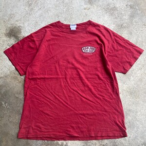 Puede incluir: Una camiseta roja de manga corta con cuello redondo y un peque&ntilde;o gr&aacute;fico en la parte superior izquierda del pecho. El gr&aacute;fico es un parche ovalado blanco y negro con el texto Ron Jon Surf Shop y Cocoa Beach, Fla. en una fuente estilizada.