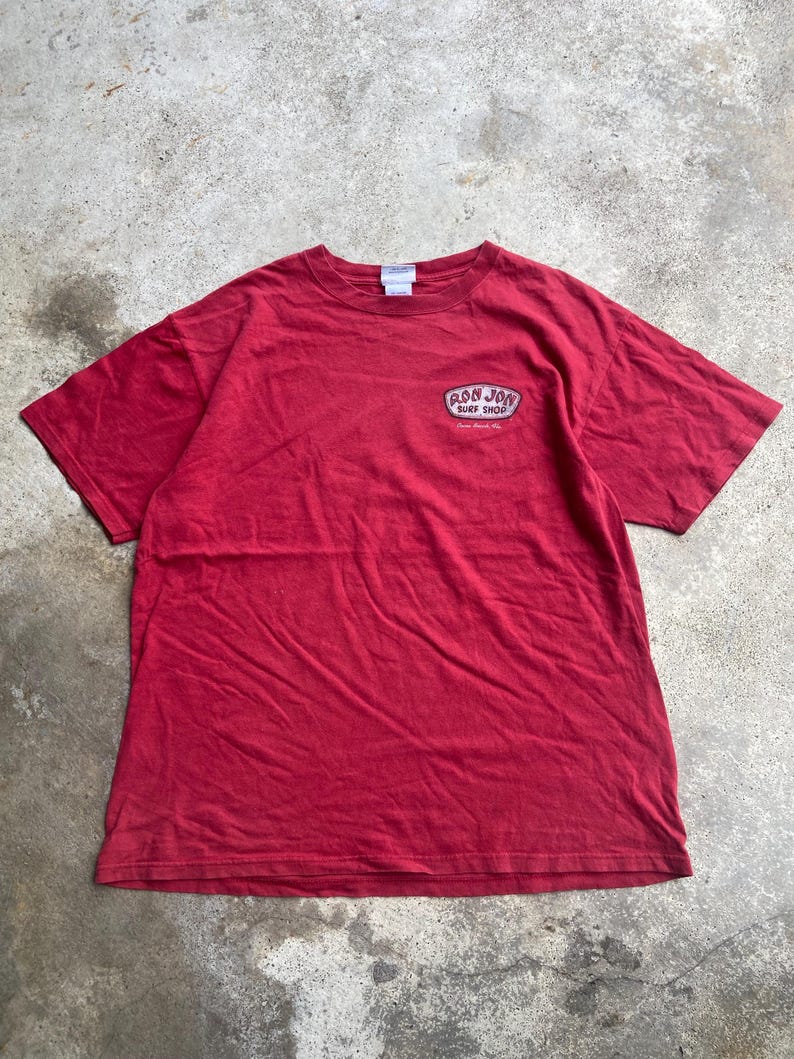 Puede incluir: Una camiseta roja de manga corta con cuello redondo y un peque&ntilde;o gr&aacute;fico en la parte superior izquierda del pecho. El gr&aacute;fico es un parche ovalado blanco y negro con el texto Ron Jon Surf Shop y Cocoa Beach, Fla. en una fuente estilizada.