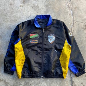 Könnte beinhalten: Eine schwarze, blaue und gelbe Reißverschlussjacke mit den Schriftzügen Rally Club London, Hardy Amies und H. Amies London Morocco Dream Rally auf der Brust. Die Jacke hat einen blauen Kragen, gelbe Seitenteile und blaue Akzente an den Ärmeln.