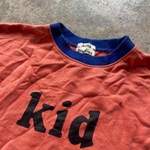 K&ouml;nnte beinhalten: Ein korallenfarbenes Sweatshirt mit einem kontrastierenden blauen gerippten Rundhalsausschnitt und dem Wort "Kid" in gro&szlig;en schwarzen Buchstaben auf der Vorderseite. Ein wei&szlig;es Innenetikett zeigt den Schriftzug "Acne Studios" in Schwarz am Kragen.
