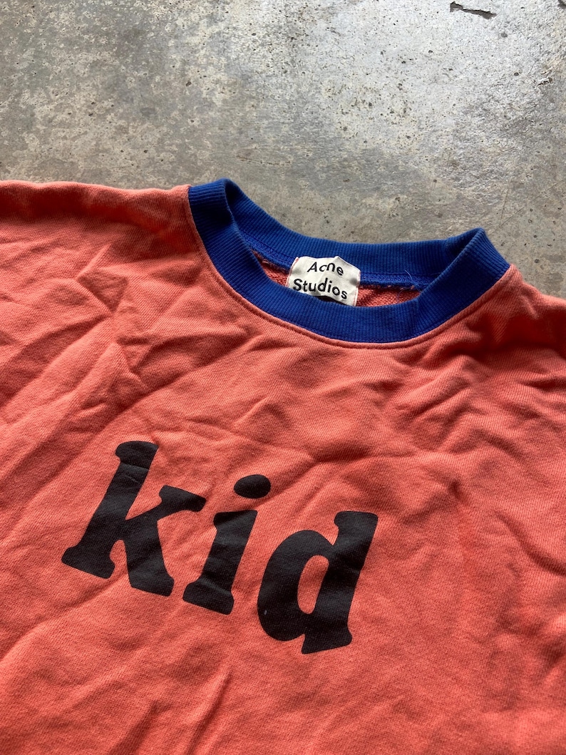 K&ouml;nnte beinhalten: Ein korallenfarbenes Sweatshirt mit einem kontrastierenden blauen gerippten Rundhalsausschnitt und dem Wort "Kid" in gro&szlig;en schwarzen Buchstaben auf der Vorderseite. Ein wei&szlig;es Innenetikett zeigt den Schriftzug "Acne Studios" in Schwarz am Kragen.