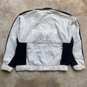 以下が含まれることがあります： 白でTHRASHERの文字がプリントされ、その下に五芒星とヤギの頭のグラフィックが描かれた、ライトグレーと黒のウィンドブレーカー。このジャケットは、黒いサイドパネルが付いた長袖、襟、伸縮性のある裾と袖口が特徴です。