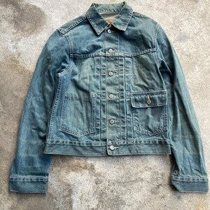 Könnte beinhalten: Eine verwaschene blaue Jeansjacke mit spitzem Kragen, langen Ärmeln mit geknöpften Manschetten und einer Knopfleiste vorne. Die Jacke verfügt über zwei vertikal gesteppte Bahnen auf der Vorderseite, eine Brusttasche mit Knopfklappe und einen geraden Saum.