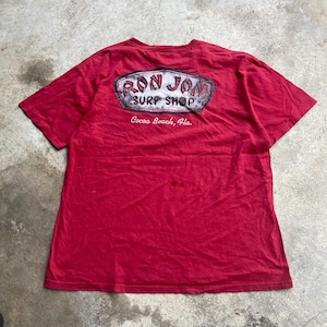 Puede incluir: Una camiseta roja de manga corta con un gr&aacute;fico en la espalda que incluye el texto "RON JON SURF SHOP" en un marco ovalado desgastado, con la ubicaci&oacute;n "Cocoa Beach, Fla." impresa en letras cursivas blancas bajo el logo.