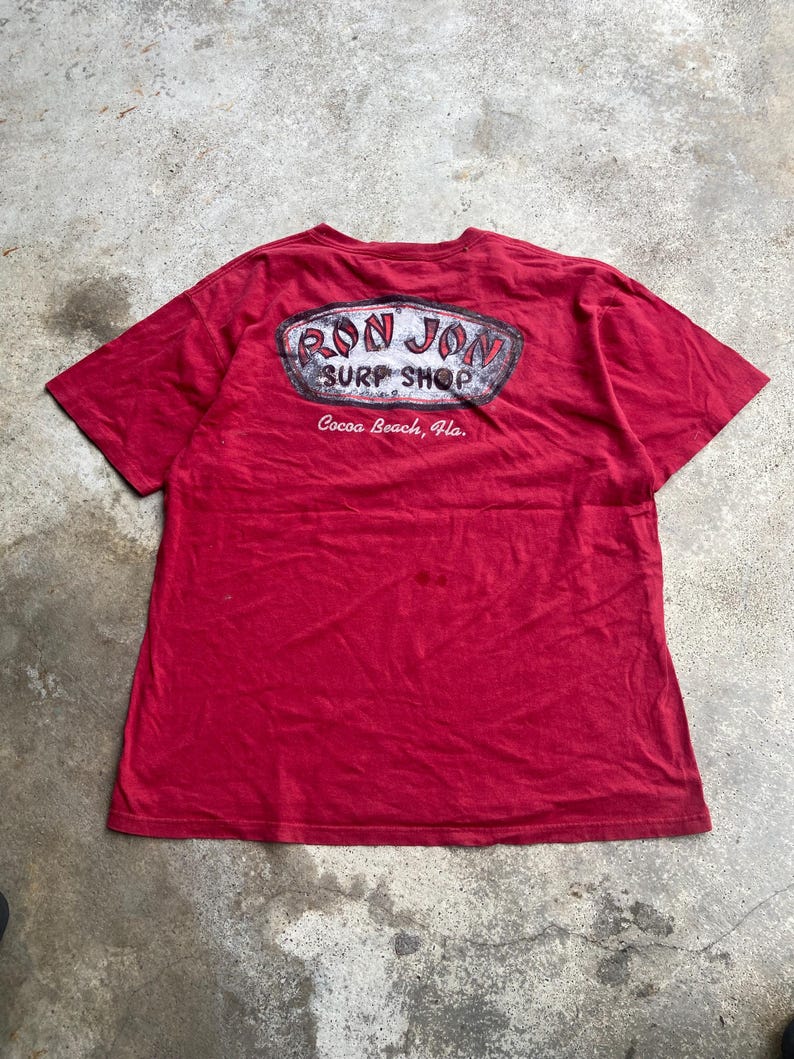 Puede incluir: Una camiseta roja de manga corta con un gr&aacute;fico en la espalda que incluye el texto "RON JON SURF SHOP" en un marco ovalado desgastado, con la ubicaci&oacute;n "Cocoa Beach, Fla." impresa en letras cursivas blancas bajo el logo.