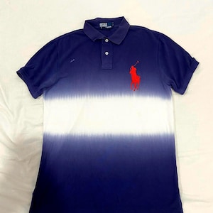 May include: A short-sleeved polo shirt featuring a dark blue and white horizontal ombre gradient pattern. The shirt has a collar, a two-button placket, and a red embroidered logo of a horse and rider on the left chest. The inner neck label reads Polo by Ralph Lauren.