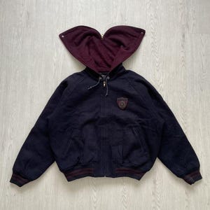 以下が含まれることがあります： ネイビーブルーのジップアップジャケット、バーガンディのハート型フード付き。 ジャケットの胸には小さなエンブレムがあり、袖口と裾にはバーガンディとネイビーのストライプが入っています。 ジャケットは厚手のテクスチャード加工された素材で作られています。