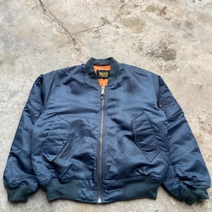 Puede incluir: Una chaqueta tipo bomber azul marino con forro interior naranja brillante, cremallera central color latón, dos bolsillos delanteros con botón a presión, un bolsillo utilitario en la manga izquierda y puños y dobladillo de punto acanalado.