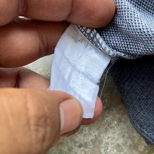 May include: Close-up of a white fabric tag with Japanese text, attached to a gray and blue patterned garment. The tag appears to have information about the garment's composition.