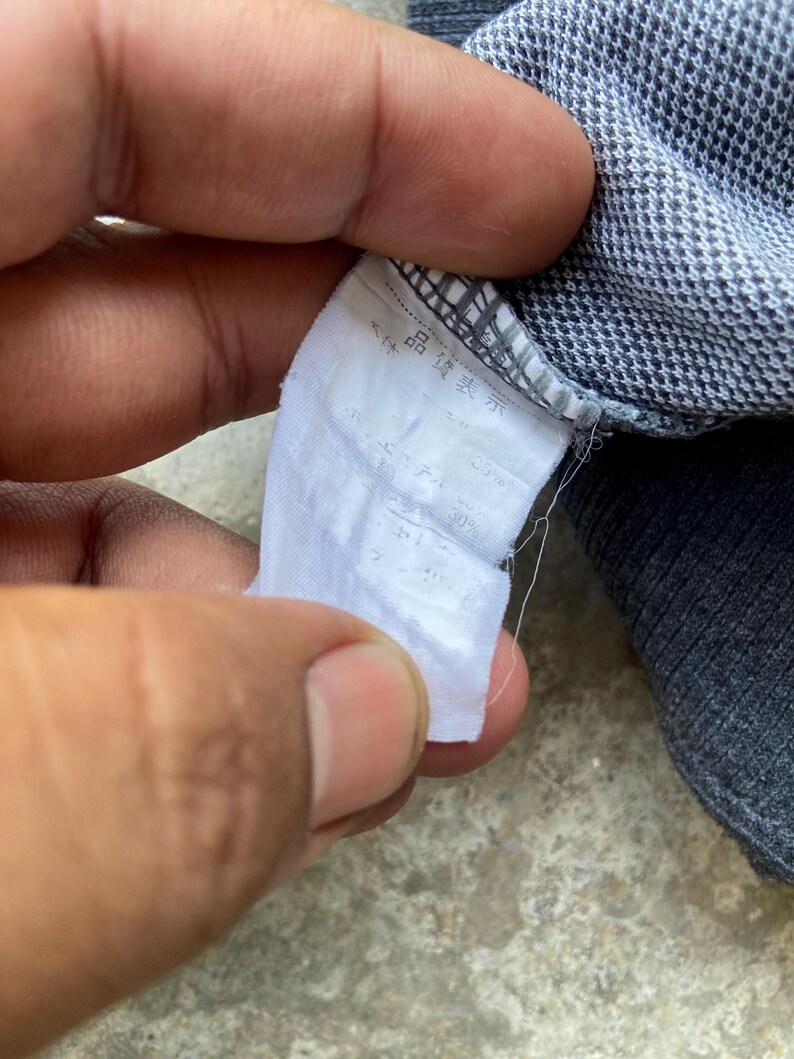 May include: Close-up of a white fabric tag with Japanese text, attached to a gray and blue patterned garment. The tag appears to have information about the garment's composition.