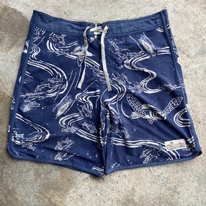 May include: Navy blue board shorts featuring a white line-art print of fish, sea turtles, and flowing water patterns. The shorts have a drawstring waist with light-colored laces and a small rectangular Pataloha brand patch on the lower right leg.