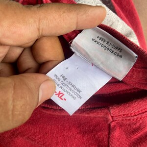 Puede incluir: Una camiseta roja con una etiqueta interior blanca. La etiqueta muestra el texto 1-888-RJ-SURFS, www.ronjons.com, PRE-SHRUNK, 100% COTTON/COTON, MADE IN HONDURAS y XL. La tela de la camiseta es de color rojo s&oacute;lido con un gr&aacute;fico gris parcial visible.