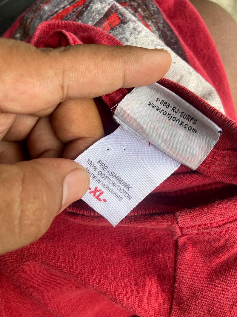 Puede incluir: Una camiseta roja con una etiqueta interior blanca. La etiqueta muestra el texto 1-888-RJ-SURFS, www.ronjons.com, PRE-SHRUNK, 100% COTTON/COTON, MADE IN HONDURAS y XL. La tela de la camiseta es de color rojo s&oacute;lido con un gr&aacute;fico gris parcial visible.