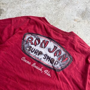 Puede incluir: Una camiseta de color granate con un gr&aacute;fico en la espalda que muestra el texto RON JON SURF SHOP en letras rojas y negras dentro de un borde ovalado desgastado, con el texto Cocoa Beach, Fla. impreso en blanco debajo.