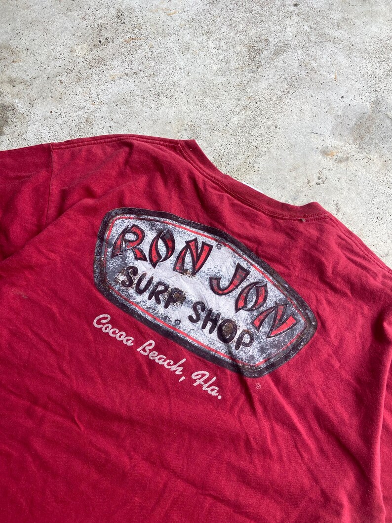 Puede incluir: Una camiseta de color granate con un gr&aacute;fico en la espalda que muestra el texto RON JON SURF SHOP en letras rojas y negras dentro de un borde ovalado desgastado, con el texto Cocoa Beach, Fla. impreso en blanco debajo.