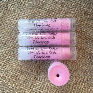 May include: Three tubes of Flamingo tinted lip balm with 10% zinc oxide. The lip balms are in clear tubes with white caps and pink balm. The text on the label includes the ingredients: organic cocoa butter, coconut oil, and beeswax.