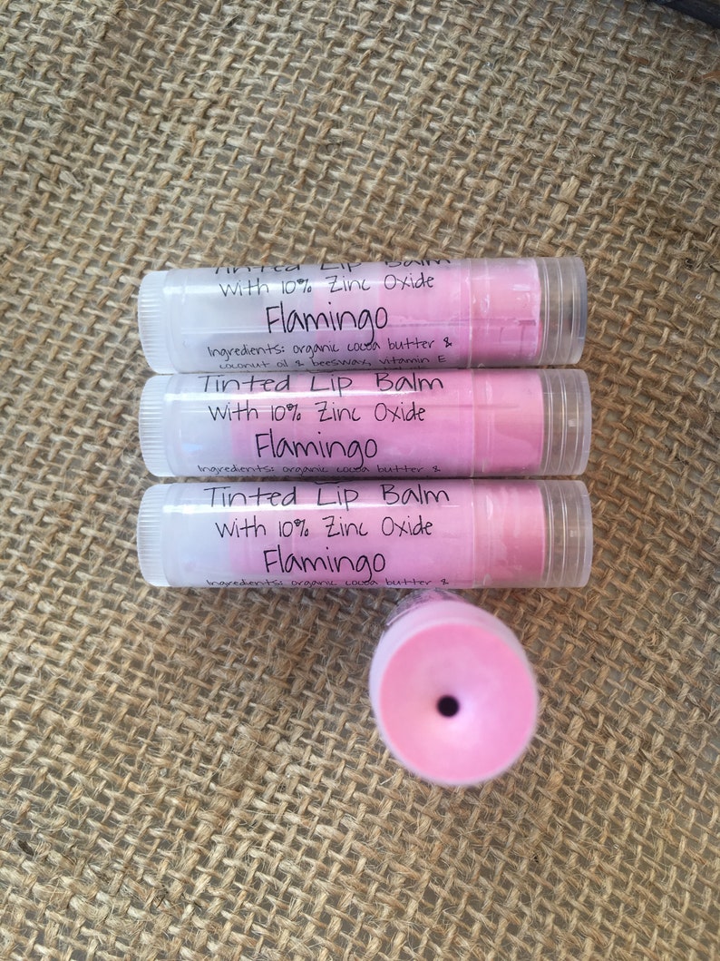 May include: Three tubes of Flamingo tinted lip balm with 10% zinc oxide. The lip balms are in clear tubes with white caps and pink balm. The text on the label includes the ingredients: organic cocoa butter, coconut oil, and beeswax.