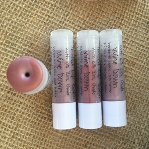 May include: Four lip balm tubes with white bases and clear tops, one with the cap off. The tubes are labeled "Wine Down Tinted Lip Balm with 10% Zinc Oxide." The lip balm is a reddish-pink color.