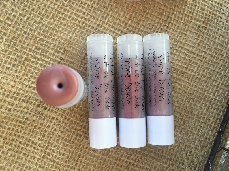 May include: Four lip balm tubes with white bases and clear tops, one with the cap off. The tubes are labeled "Wine Down Tinted Lip Balm with 10% Zinc Oxide." The lip balm is a reddish-pink color.
