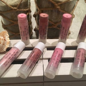 May include: Lip balms in various shades of pink and red are displayed on a white surface. The lip balms are in clear tubes with white caps. The text on the tubes includes "With Zinc Oxide" and flavor names. Seashells and decorative jars are in the background.