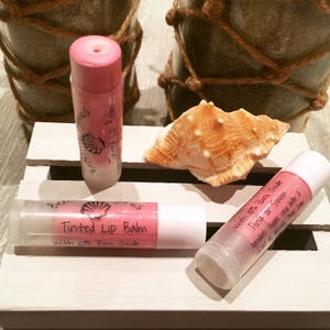 May include: Three lip balm tubes with pink tinted balm and white caps are displayed. The tubes have labels that read "Tinted Lip Balm" and "With 10% Zinc Oxide." A seashell and decorative jars are in the background.