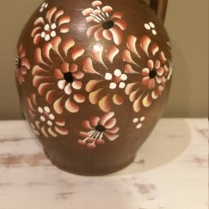 May include: A brown ceramic pitcher with a handle, painted with white and pink flowers. The flowers have black centers.