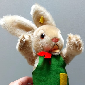 Op de afbeelding: Een pluche bruin konijn met een groene schort met een gele zak en een rode strik. Het konijn heeft een geel label op zijn oor.