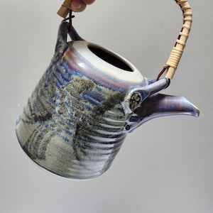 May include: A ceramic teapot with a woven handle. The teapot has a blue, purple, and gray glaze with a textured surface. The handle is made of woven bamboo. The spout is a dark purple color.