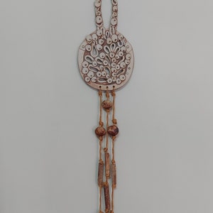 May include: A ceramic wall hanging with a circular design and a brown and white colour scheme. The hanging features a loop at the top and a long string of brown beads and wooden dowels hanging from the bottom.
