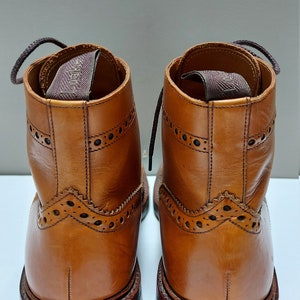 May include: A pair of brown leather lace-up boots with brogue detailing. The boots have a brown leather sole and a brown leather heel.
